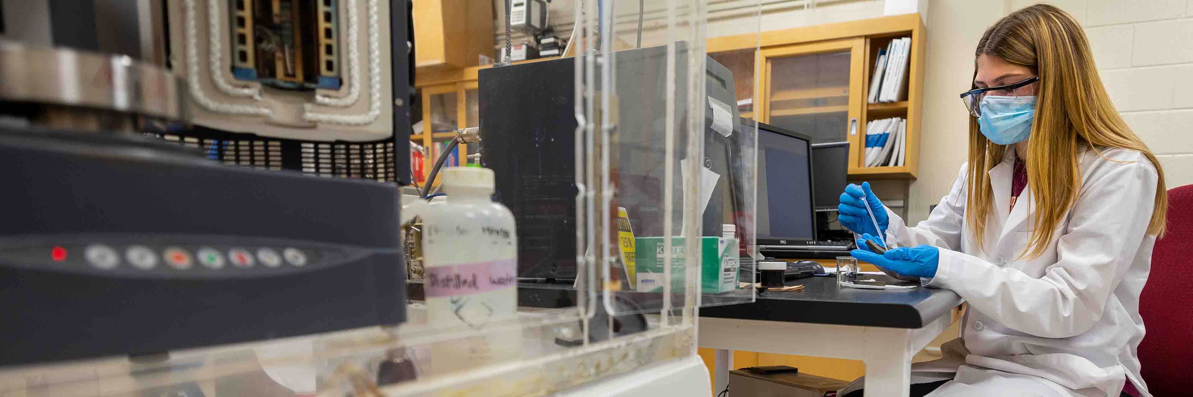 A student researcher wearing a lab coat, mask, glasses, and latex gloves uses a pipet on a sample of hemp.