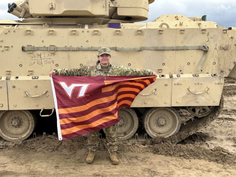 Richmond stands by a tank in a camouflage uniform holding a striped Virginia Tech flag. 