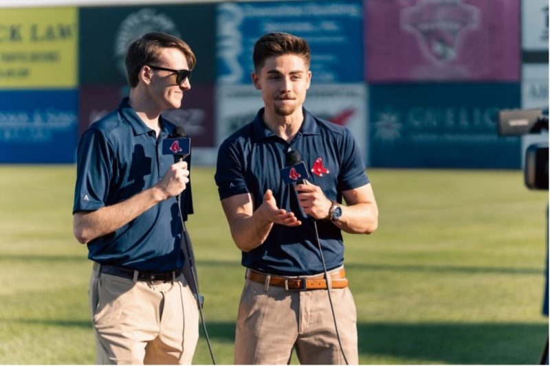 Two male students stand on a baseball field with microphones.