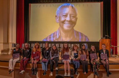 Ten students, seated on a stage, hold certificates. In the background, on a large screen, is a photo of Nikki Giovanni, smiling.