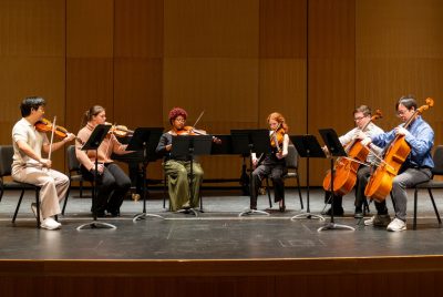Six students sit on stage, playing violins, violas, and cellos.