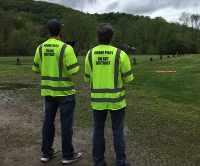 Drone pilots fly drone across green pasture on a cloudy day