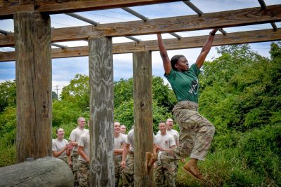 A woman cadet in a military uniform swings from a monkey bar obstacle and looks determined as she crosses it successfully. Cadets cheer in the background.