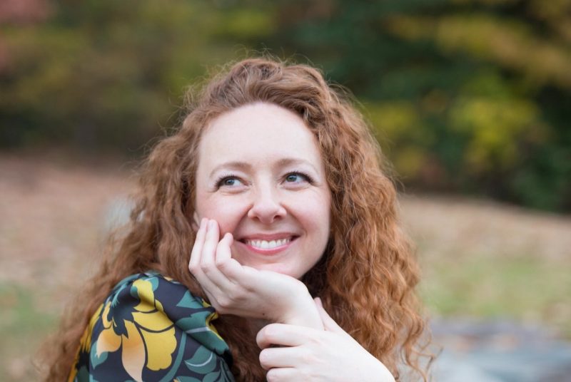 Francesca Hurst, red hair, flowered print top, leaning her chin on her hand and smiling