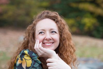 Francesca Hurst, red hair, flowered print top, leaning her chin on her hand and smiling