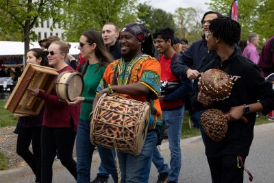 A Black man in brightly colored traditional clothing and carrying a large sideways drum walks in the middle of a group of people in more modern western-style dress. They carry various instruments that they are in the act of playing. Sheltered booths, trees, and a crowd of people can be seen in the background.