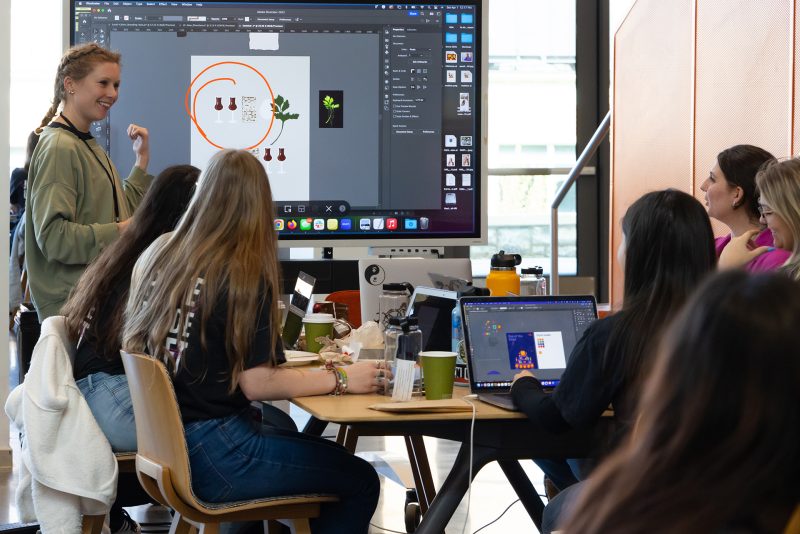 Faculty member pointing at board with students around a table listening