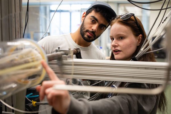 Two student researchers in the lab, the one on the right is pointing at a circular opening.